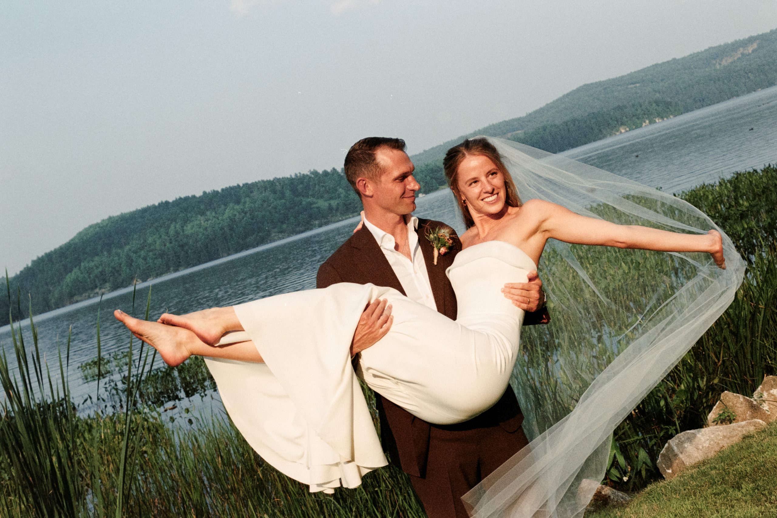 Groom carrying bride in front of waterfront at cottage